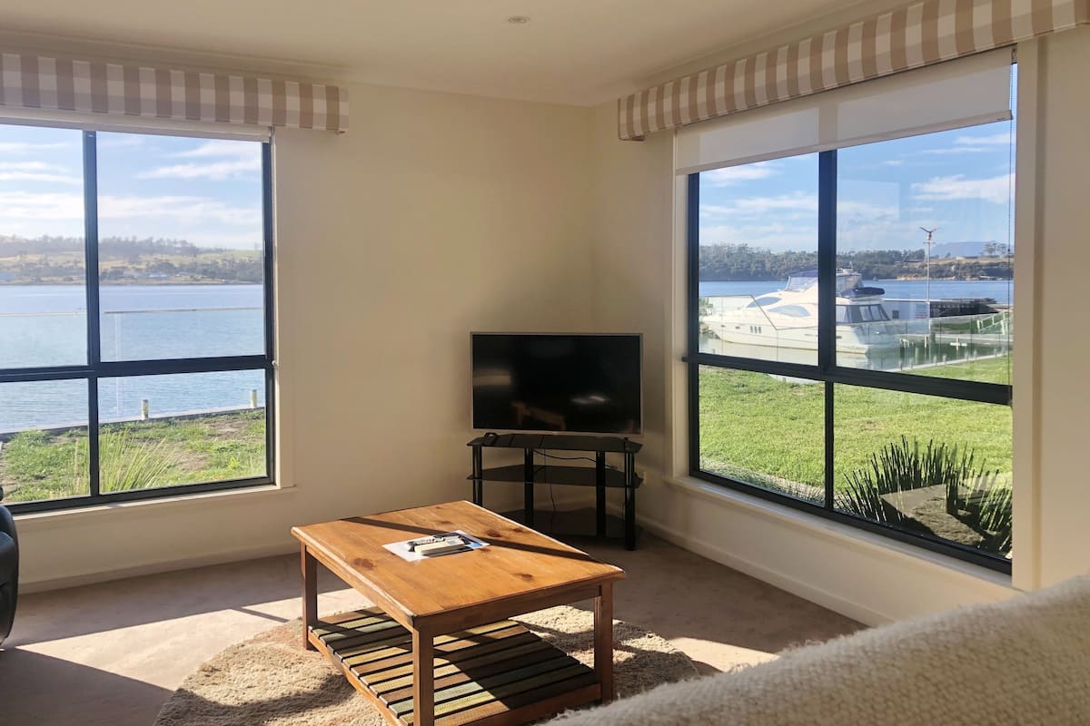 Ein Zimmer mit großem Fenster und Blick auf einen See und Boote, einem Holztisch und einem Flachbildfernseher.