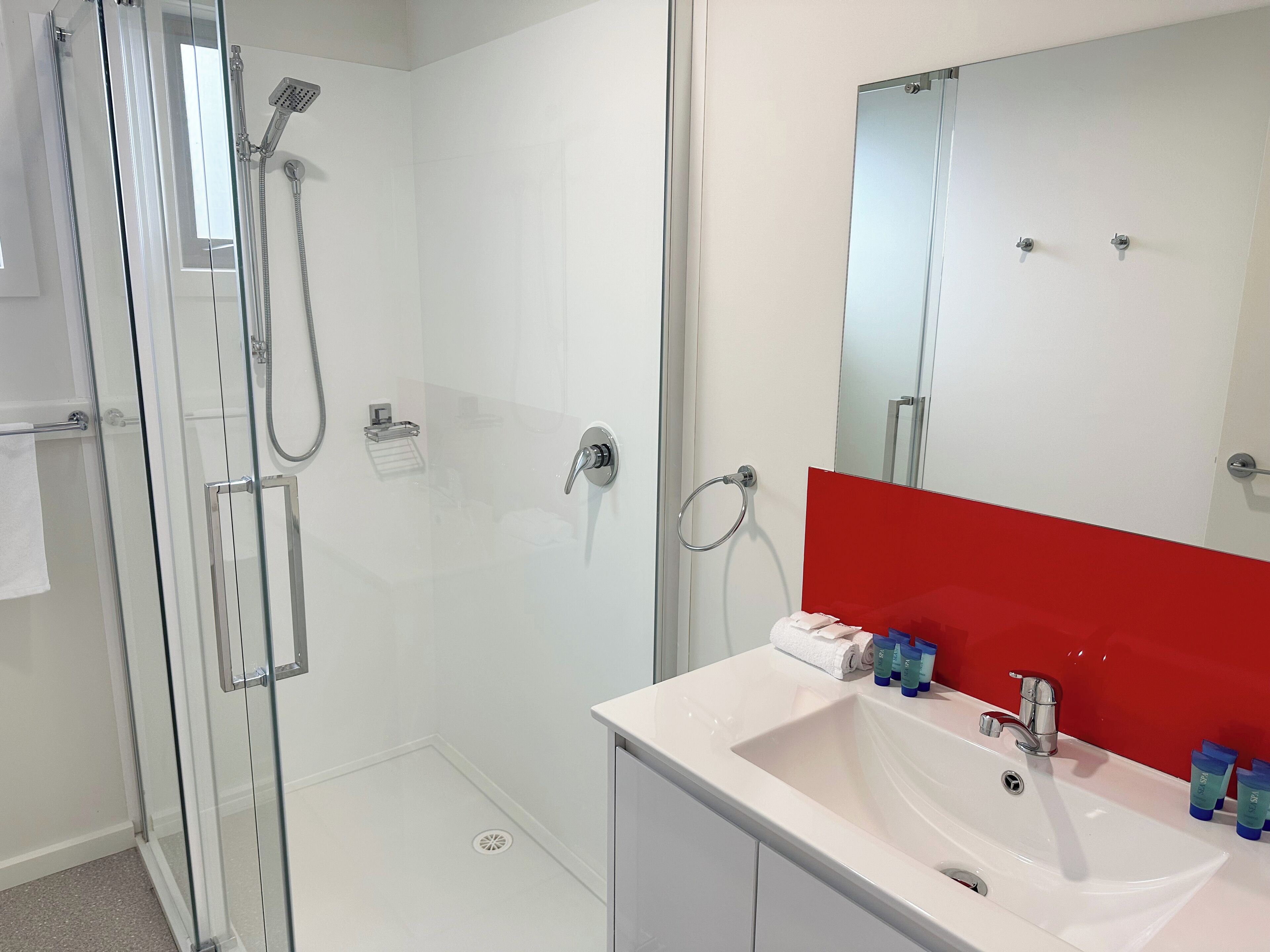 A modern bathroom with a glass-enclosed shower, a white sink with a red accent, and a large mirror.