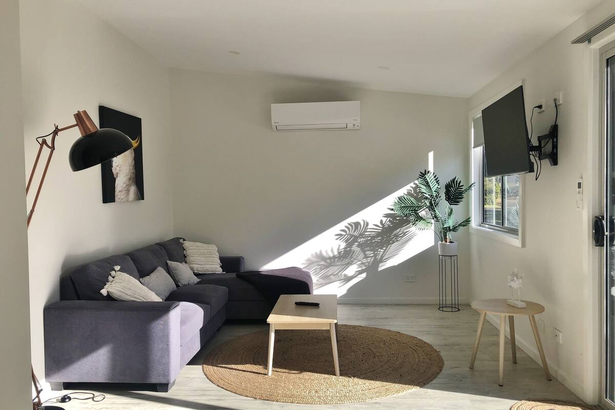 Ein modernes Wohnzimmer mit einem grauen Ecksofa, einem kleinen Holztisch, einem runden Webteppich und einem Blumentopf am Fenster.