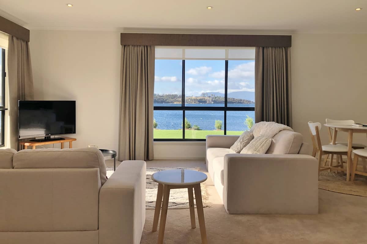 Ein modernes Wohnzimmer mit einem großen Fenster, das einen malerischen Blick auf einen See und entfernte Hügel bietet. Ein Flachbildfernseher steht auf einem Holzgestell, eine beigefarbene Couch, ein kleiner runder Tisch und ein Essbereich mit einem weißen Tisch und Holzstühlen sind ebenfalls vorhanden.