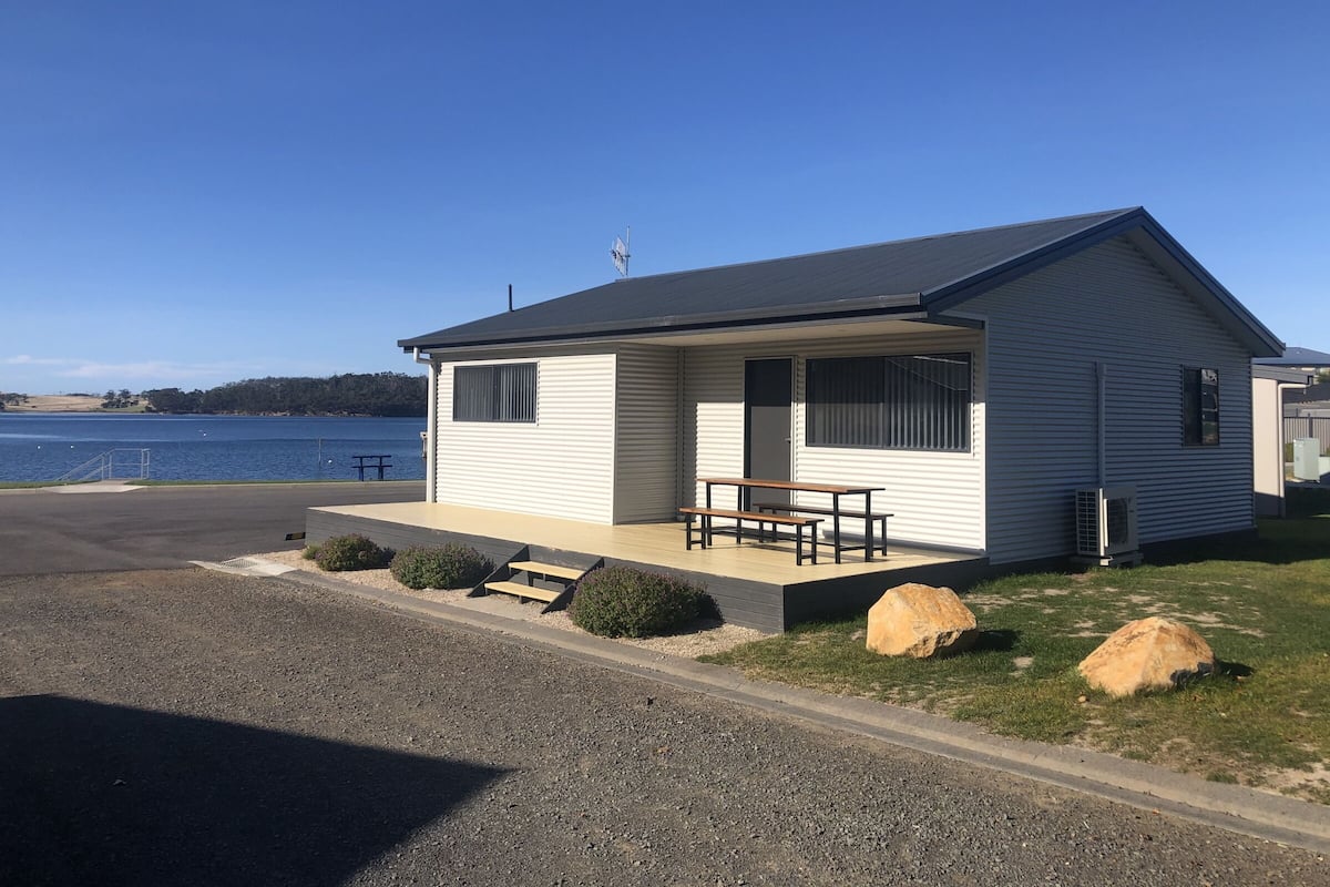 Ein Bungalow mit überdachter Veranda, einem kleinen Tisch und Blick auf einen See.