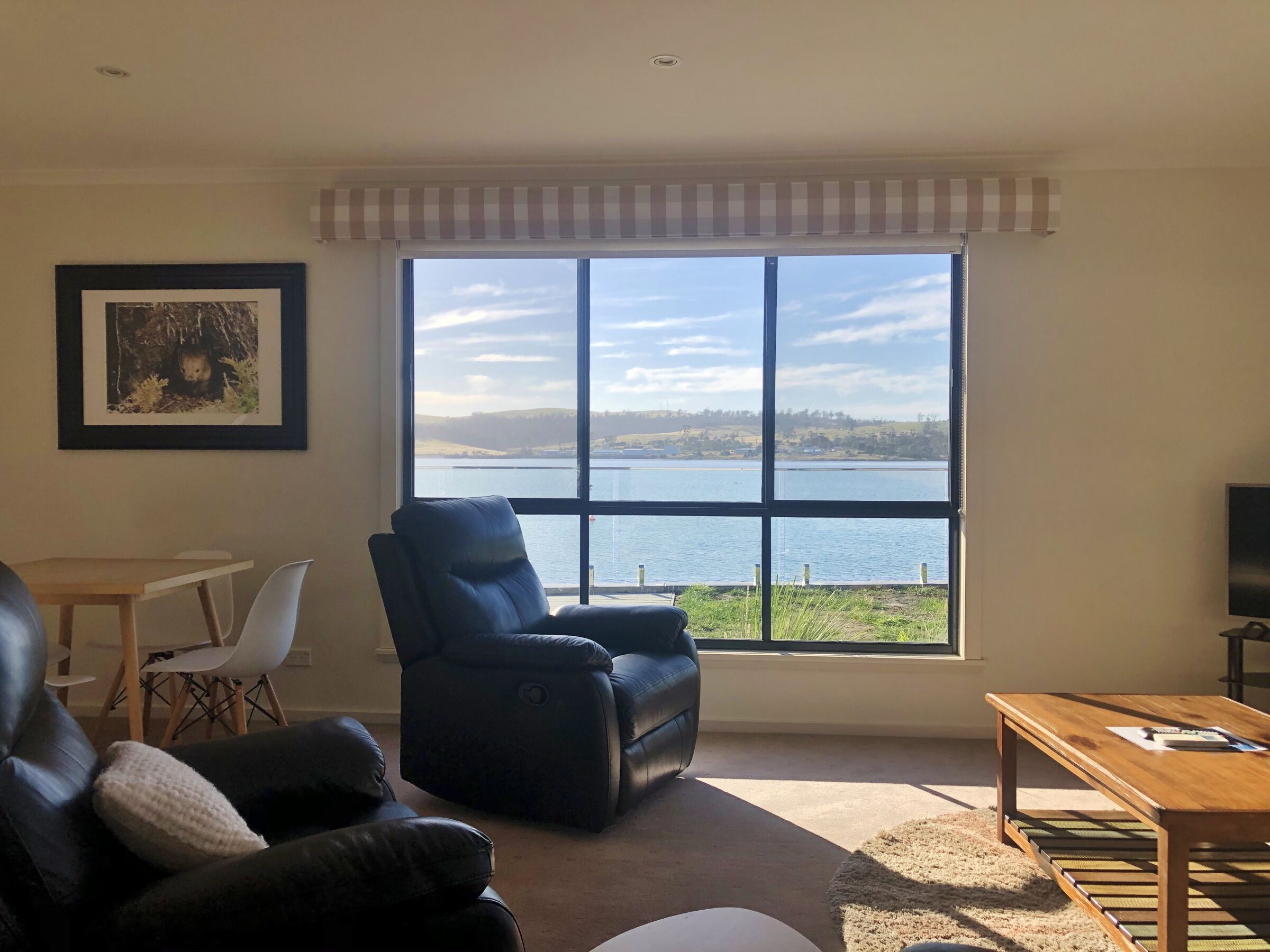Una sala de estar con un amplio ventanal que ofrece vistas a un lago y colinas. Hay un sofá de cuero negro, una mesa de centro de madera y un cuadro en la pared.