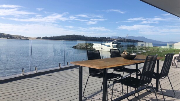 Een houten tafel met vier stoelen op een terras met uitzicht op een meer, met een jacht die er afgemeerd ligt.