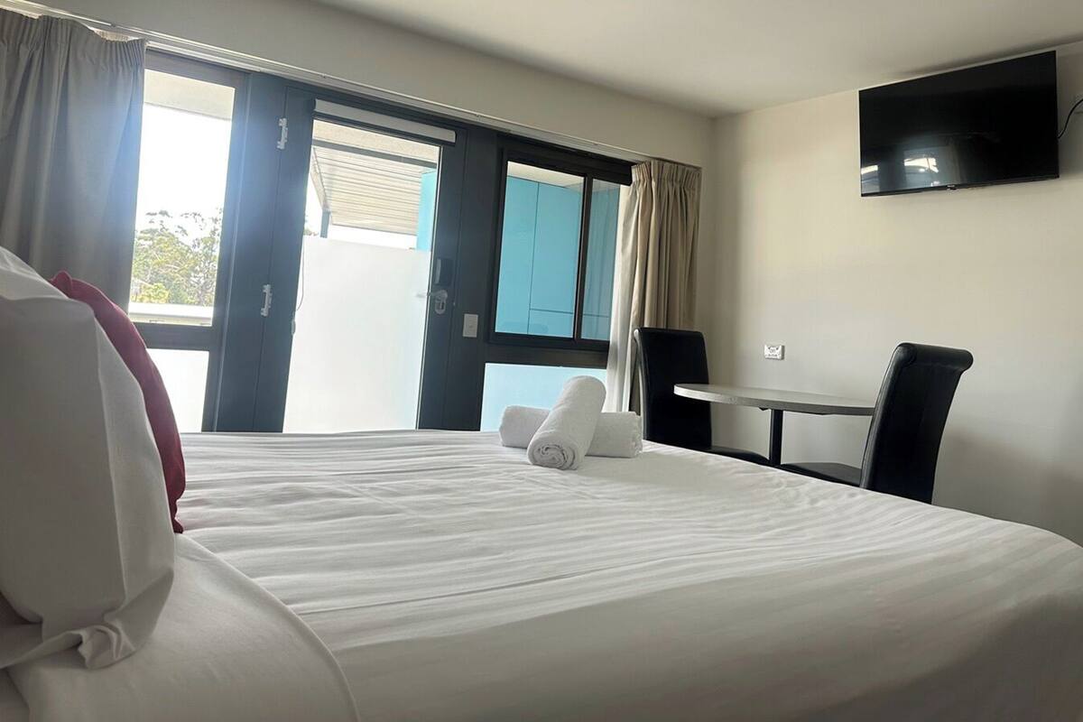 Ein Hotelzimmer mit einem großen Bett, einem Schreibtisch, einem Sessel, einem an der Wand befestigten Fernseher und einem Fenster mit Vorhängen.