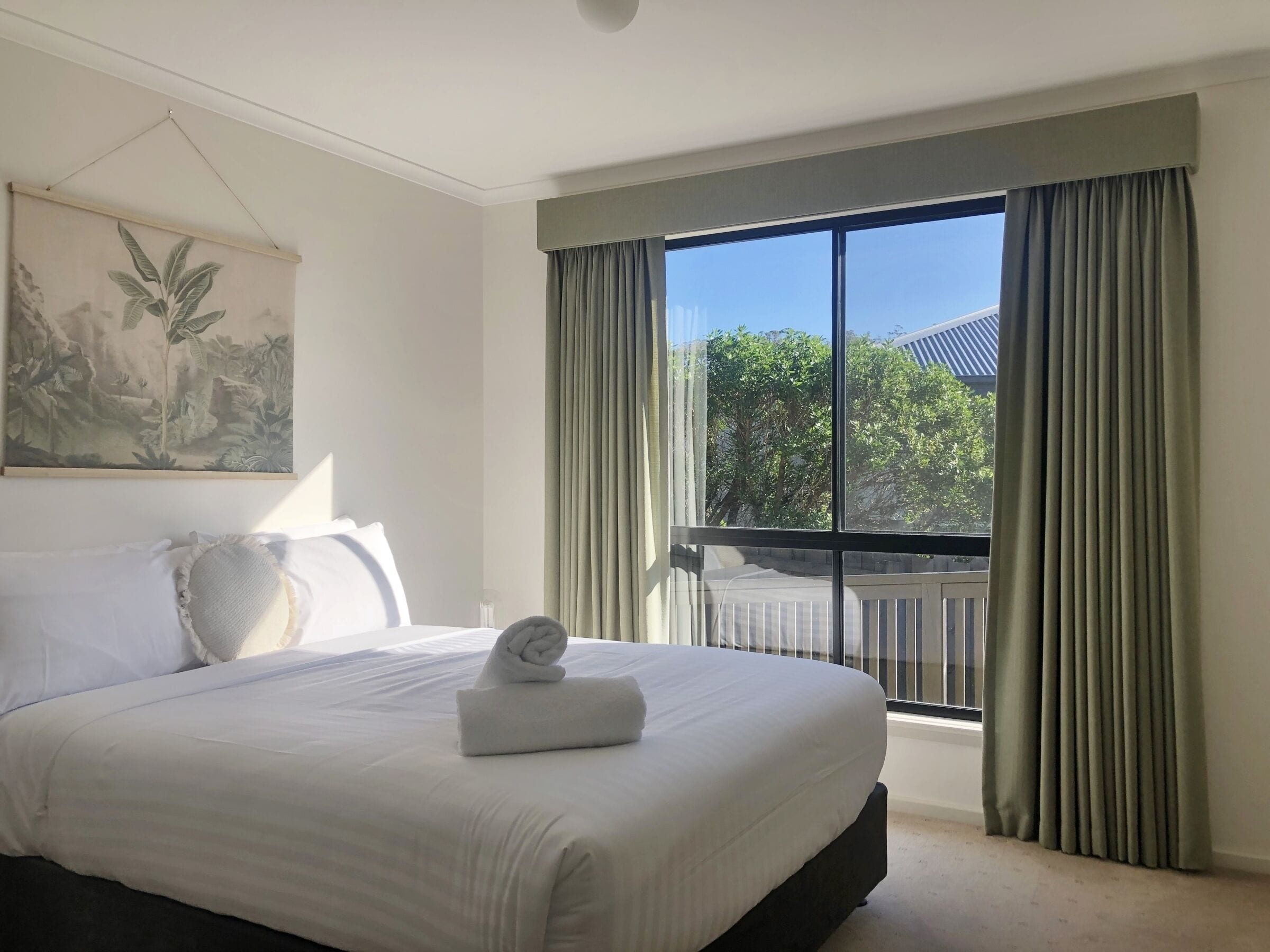 A neatly made bed with a rolled towel, a painting on the wall, and a balcony with curtains.