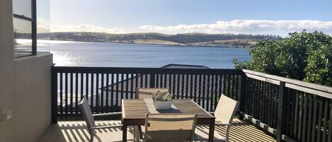 A wooden deck with a table and chairs, overlooking a body of water and distant hills.