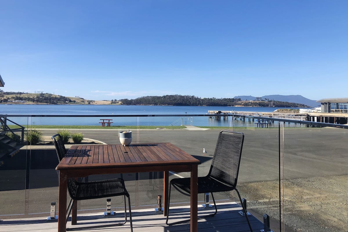Ein Holztisch und zwei Stühle auf einer Terrasse mit Blick auf einen See und einen Steg.