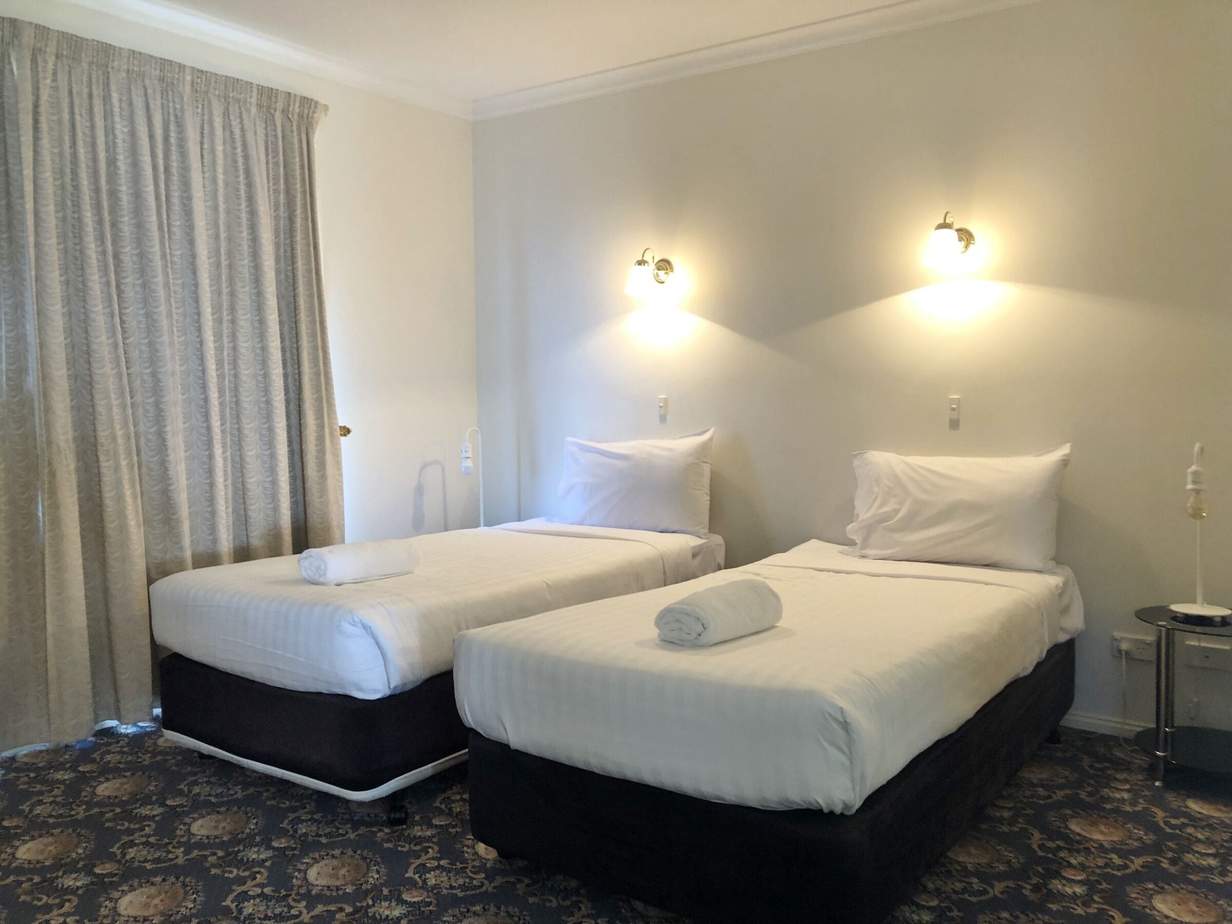 A hotel room with two beds, each with white linens and pillows, a small bedside table, and a wall-mounted light fixture.