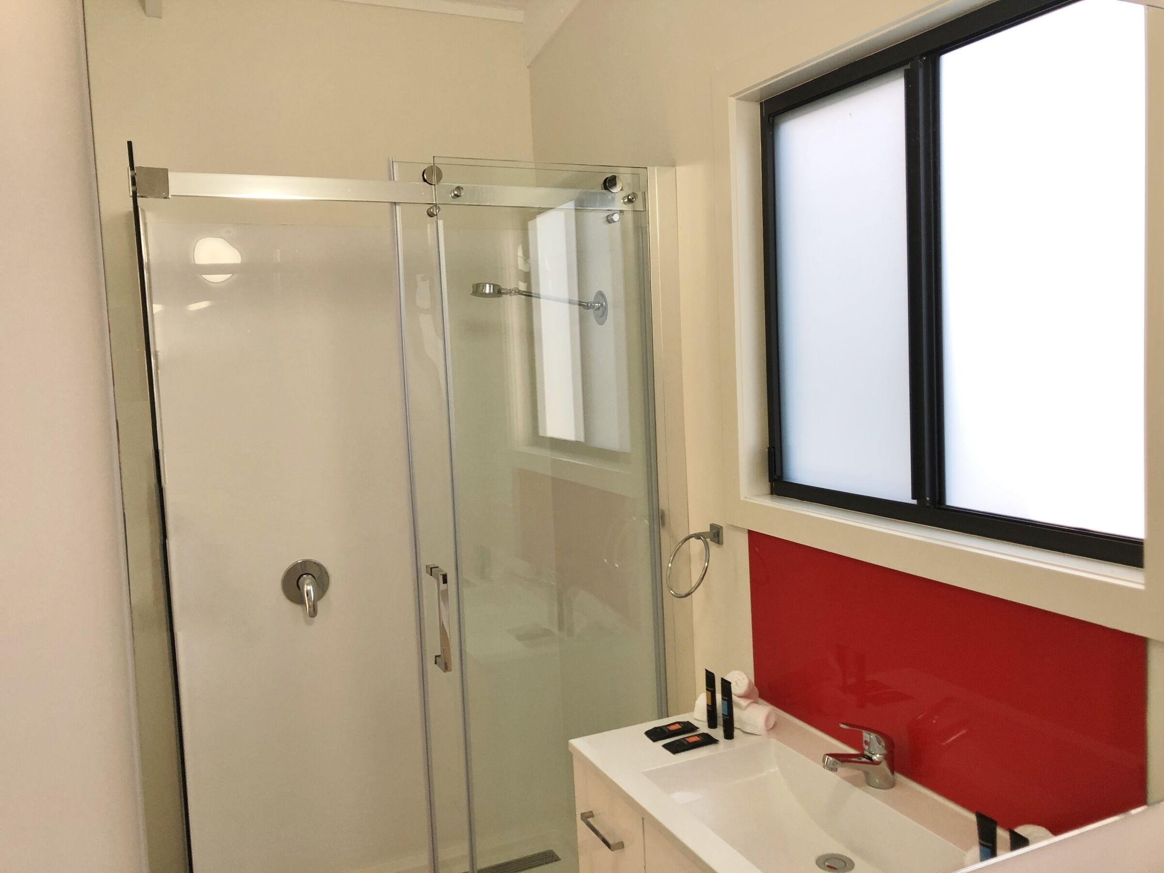 Een moderne badkamer met een glazen douchecabine, een witte wastafel en een rode accentmuur, met een raam boven de wastafel.