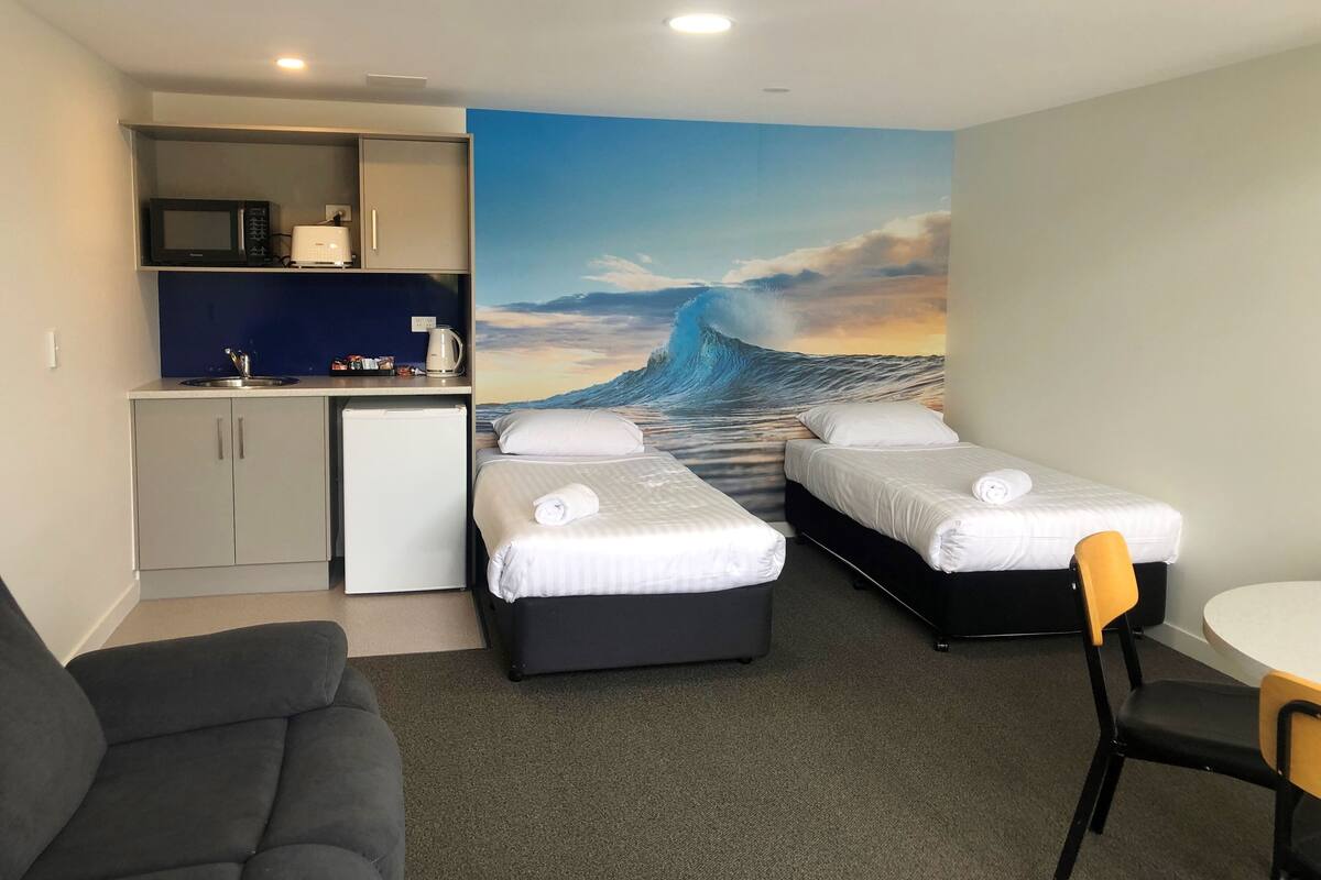 Ein kompaktes Hotelzimmer mit zwei Einzelbetten, einer kleinen Küchenzeile, einem Schlafsofa und einem Esstisch.