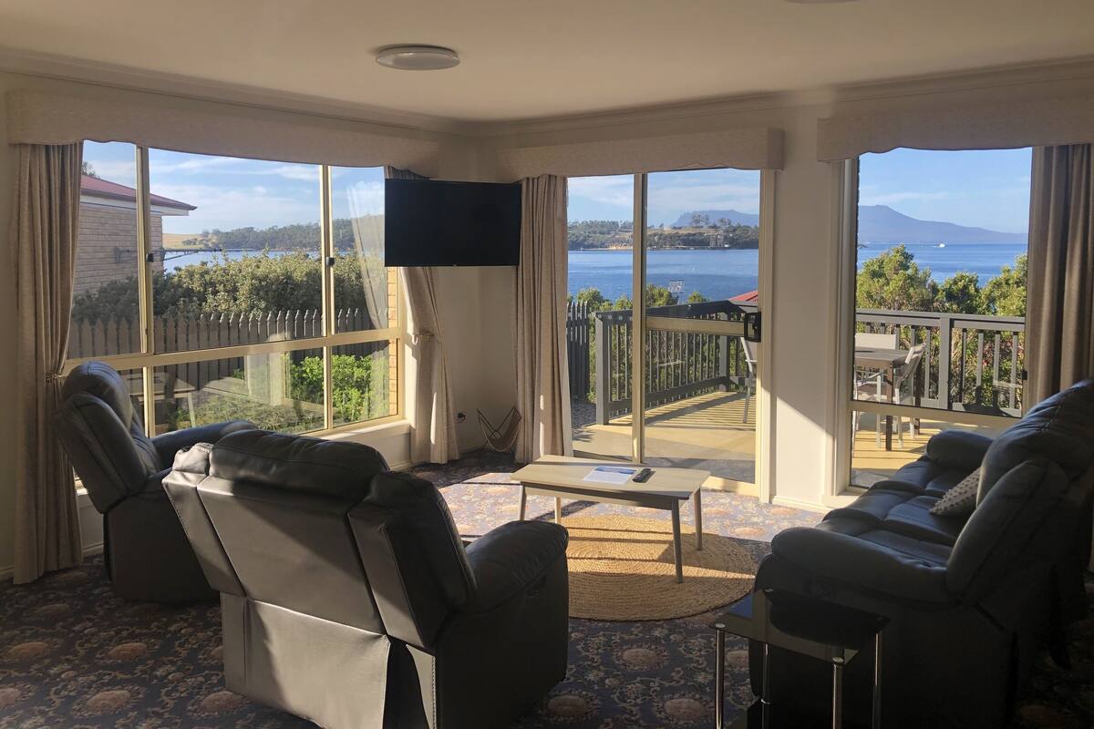 Ein Zimmer mit großem Fenster und Blick auf einen See und Berge. Zwei Liegesessel und ein kleiner Tisch sind vorhanden.