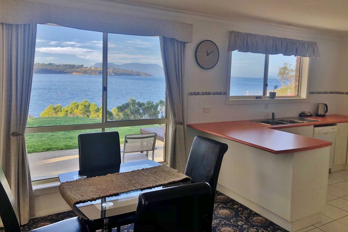 Ein Zimmer mit Esstisch, Stühlen, einer Küchenzeile mit Spüle und Arbeitsfläche sowie einem großen Fenster mit Blick auf das Meer und die Natur.