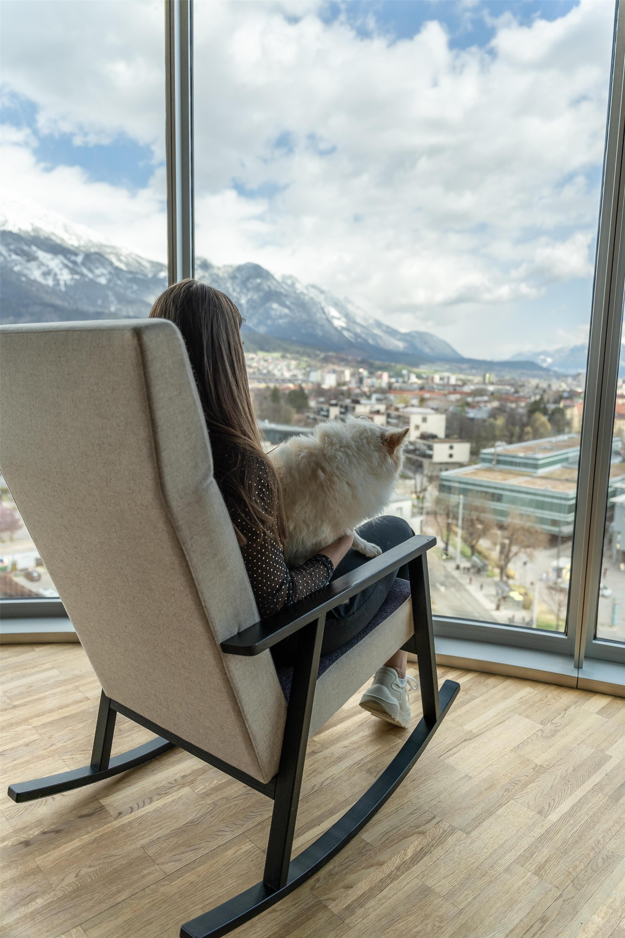 Une personne assise dans un fauteuil à bascule, un chat à ses côtés, et qui contemple un paysage urbain et des montagnes à travers de grandes fenêtres.