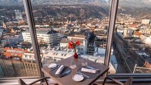 A dining area with a table set for two, a vase with flowers, and a view of a cityscape and mountains through large windows. - ADLERS Hotel (Innsbruck)