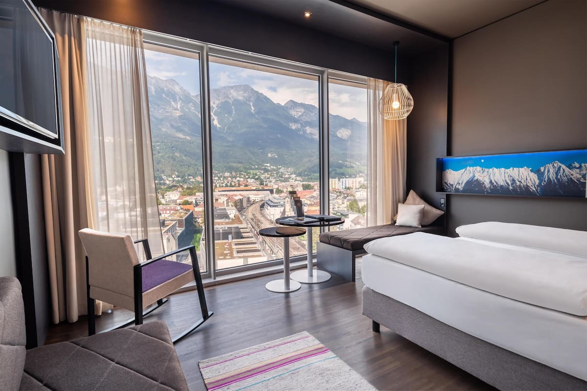 Habitació d'hotel moderna amb una finestra gran que ofereix vista a la muntanya, un sofà, una taula petita i un televisor fixat a la paret.