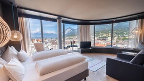 A modern hotel room with a large bed, a sofa, a TV, and a view of a cityscape and mountains. - ADLERS Hotel (Innsbruck)