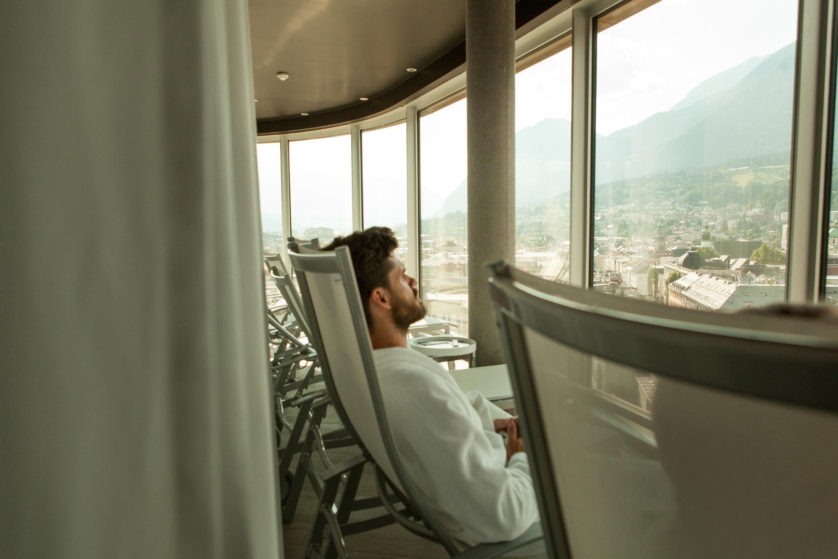 A man seated by a large window overlooking a cityscape and mountains.