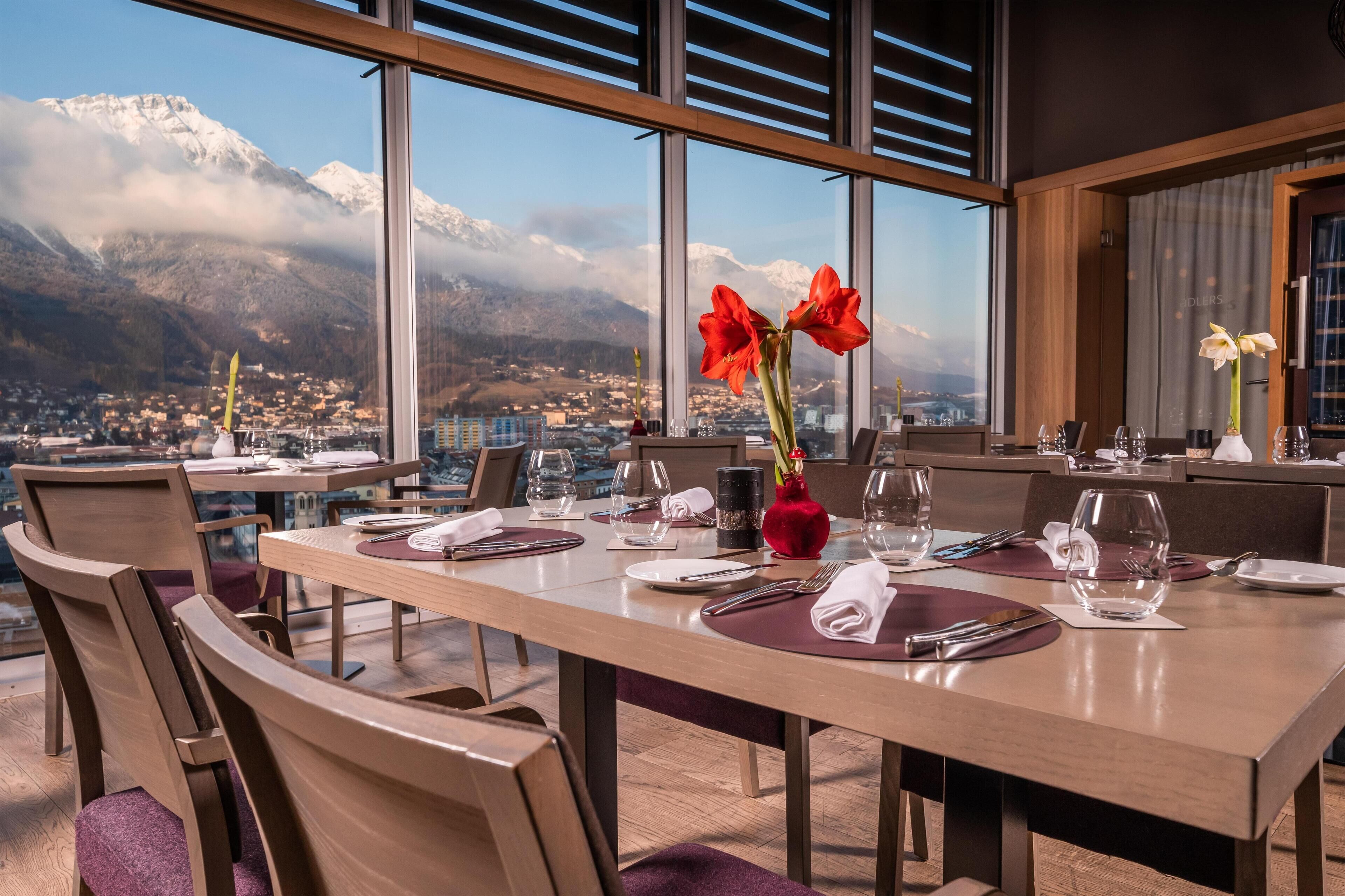 A dining area with a table set for two, large windows offering a view of snow-capped mountains and a town below.