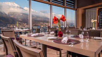 A dining area with a table set for two, large windows offering a view of snow-capped mountains and a town below.