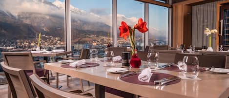 Un comedor con una mesa lista para dos, ventanas amplias que ofrecen vistas de montañas nevadas y un pueblo en la falda.