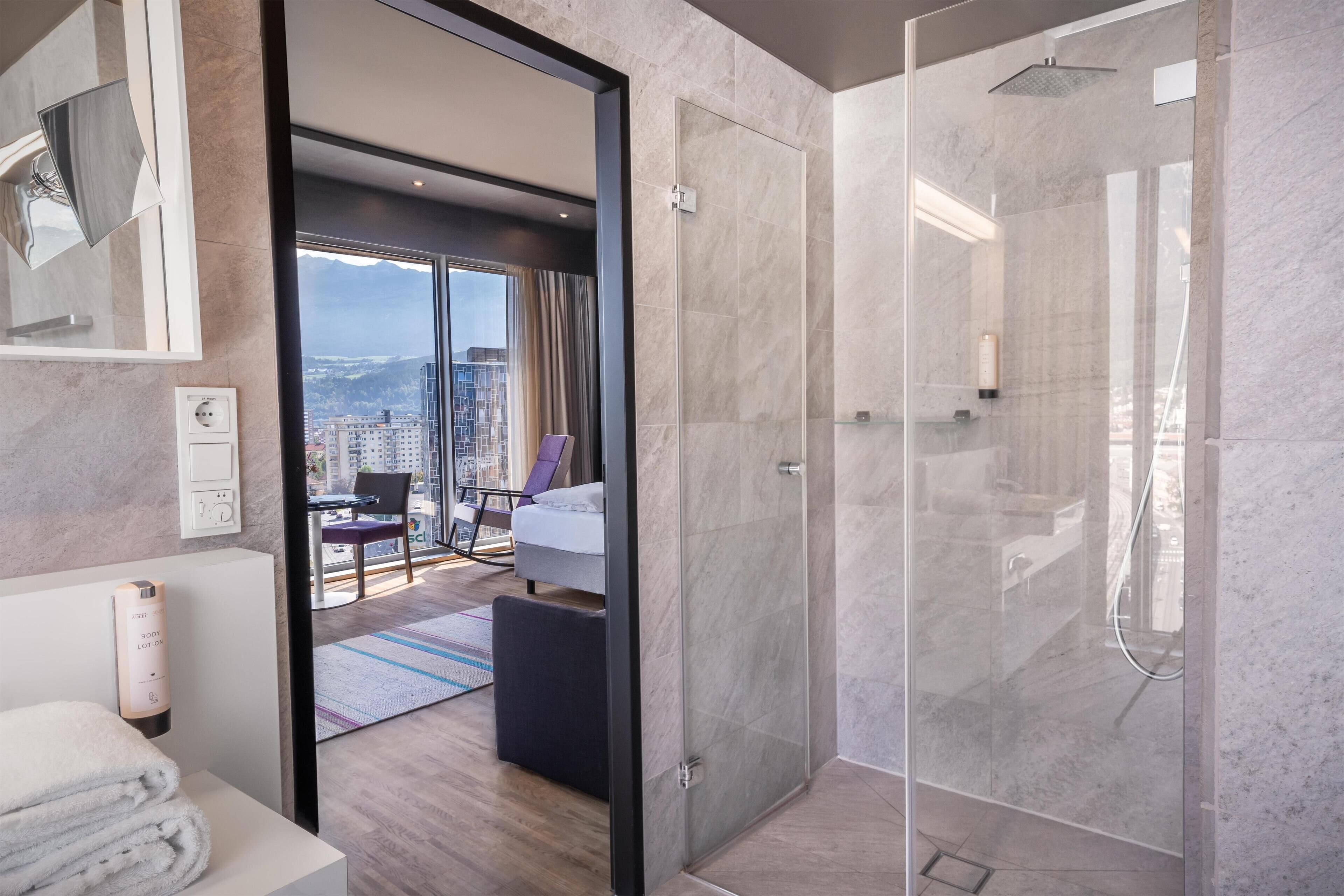 A modern hotel bathroom with a glass-enclosed shower, a visible city view, a neatly made bed, and a bathroom counter with towels.