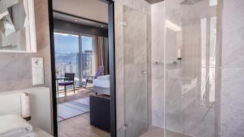 A modern hotel bathroom with a glass-enclosed shower, a visible city view, a neatly made bed, and a bathroom counter with towels.