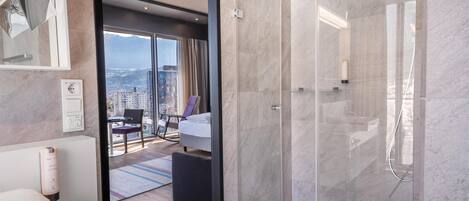 A modern hotel bathroom with a glass-enclosed shower, a visible city view, a neatly made bed, and a bathroom counter with towels.