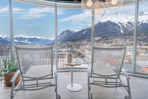 Un balcón con dos sillas y una mesa, con vistas a una ciudad y montañas nevadas.