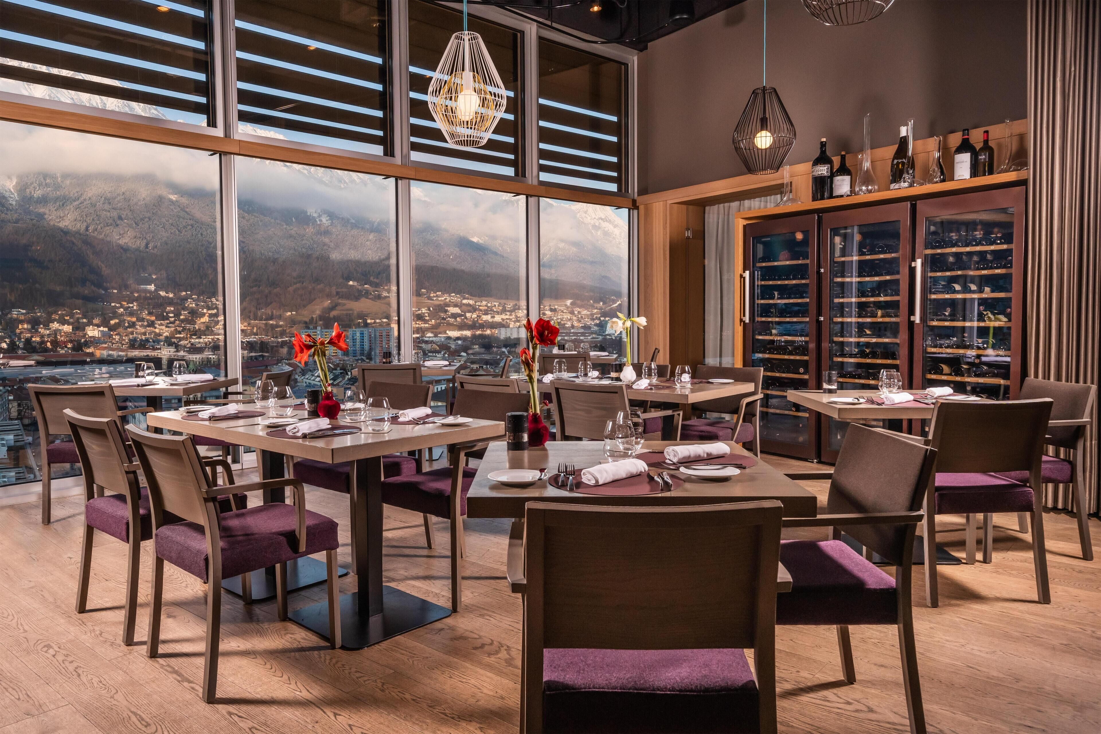 Un espace repas avec des tables disposées pour accueillir quatre personnes, de grandes fenêtres offrant une vue sur la ville et les montagnes, et un meuble vitrine pour exposer les bouteilles de vin.