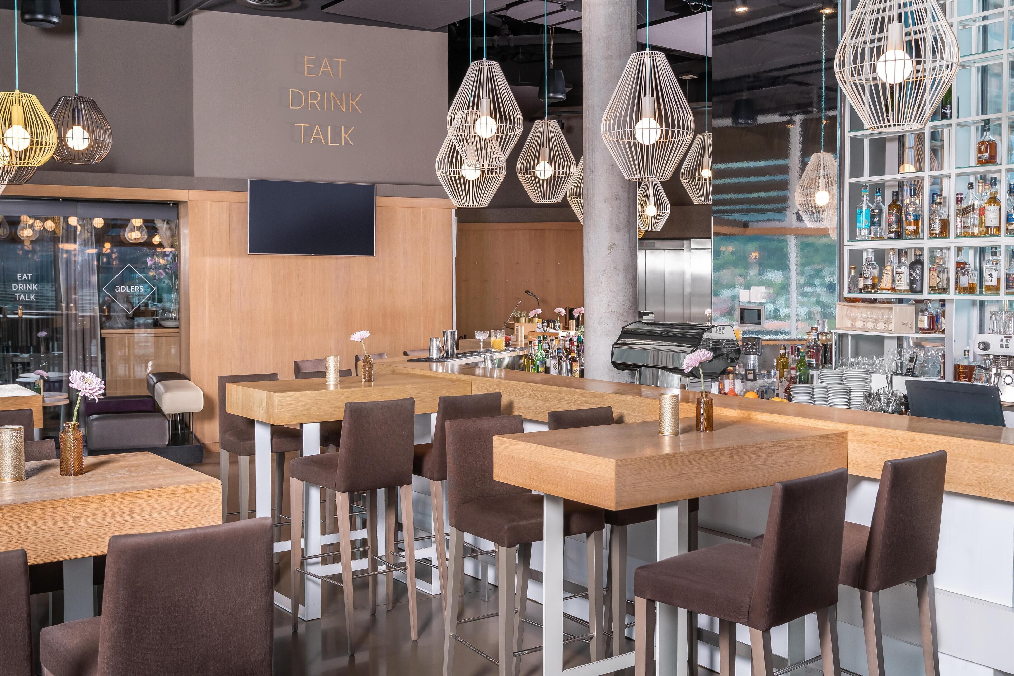 A modern bar area with wooden tables and chairs, hanging pendant lights, and a bar stocked with bottles.