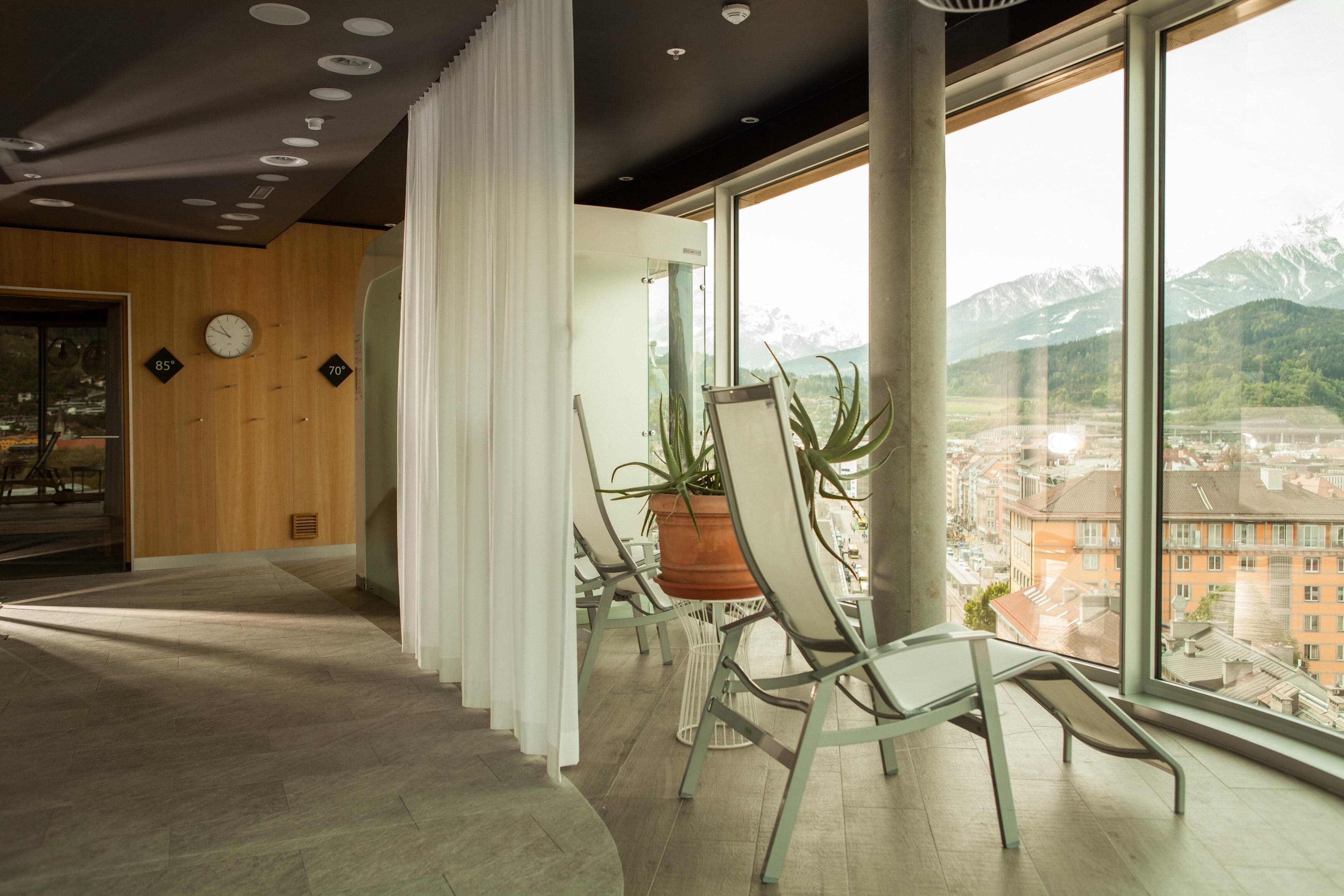A modern interior with a large window offering a mountain view, a wooden wall with a clock, and a seating area with chairs and a potted plant.