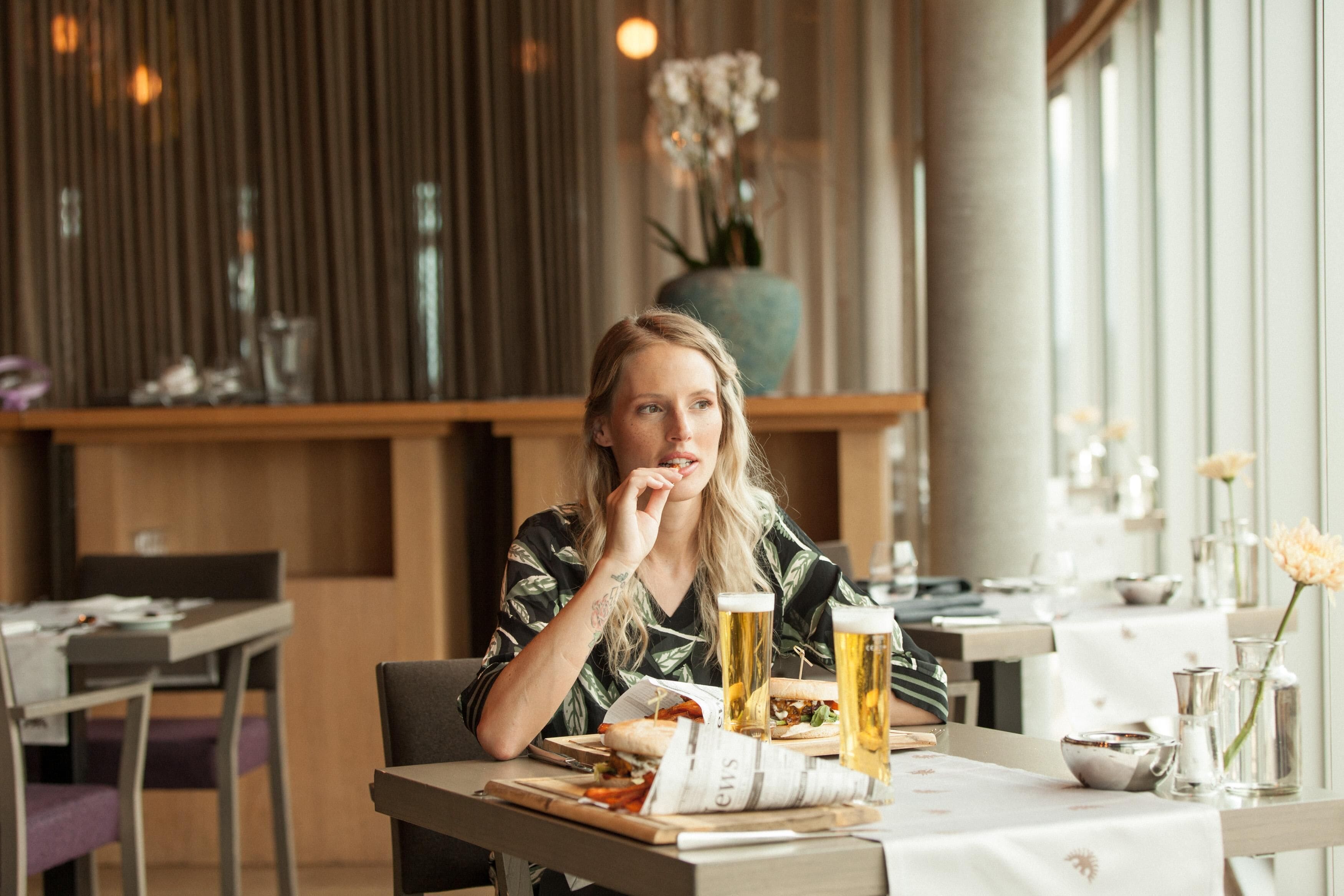 Una persona sentada en una mesa con comida y dos vasos de cerveza, en un restaurante con decoración de madera y ventanas amplias.