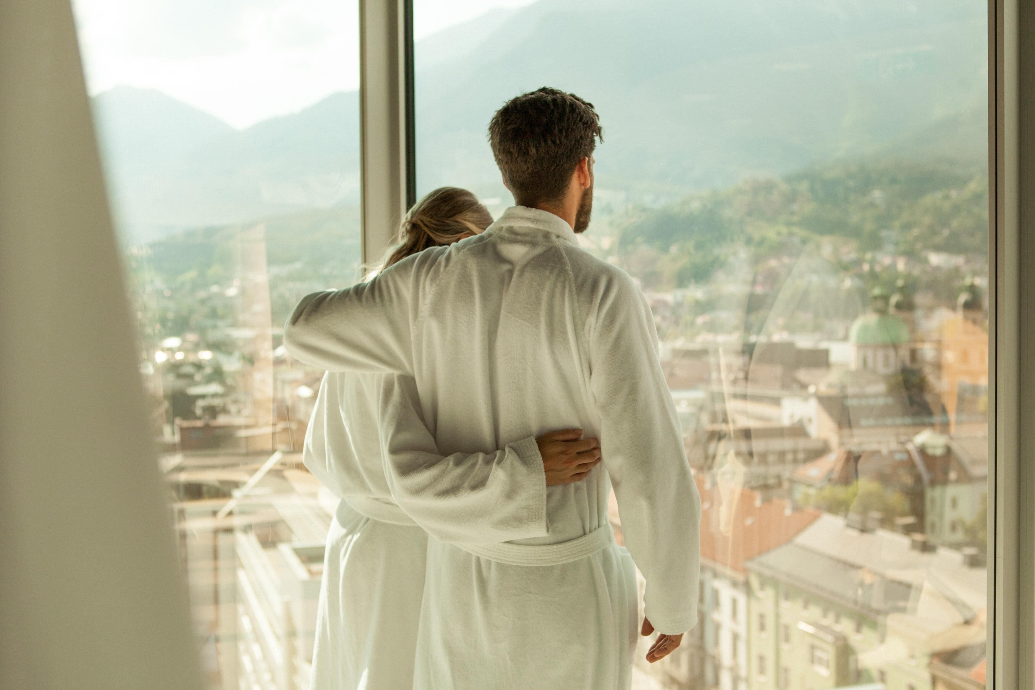 A couple embracing in a room with a large window overlooking a cityscape and mountains.