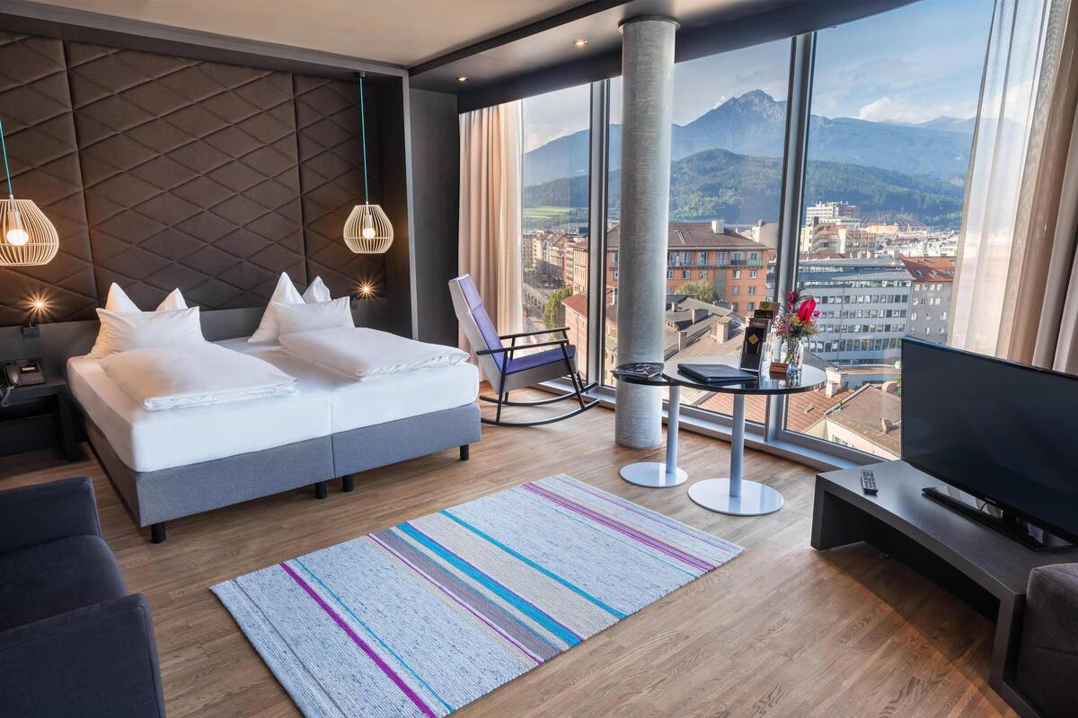 Habitació d'hotel moderna amb un llit gran, una televisió, zona de seients i vista a la ciutat i a les muntanyes.