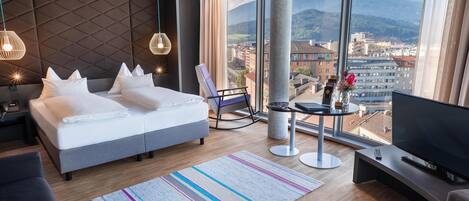 A modern hotel room with a large bed, a TV, a seating area, and a view of a cityscape and mountains.