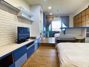 A hotel room with a TV on a white brick wall, a bed, a small table, and a window with curtains. - MB House (Kaohsiung)