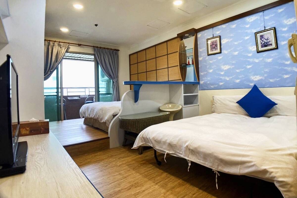 Une chambre d’hôtel avec deux lits, une petite table, une chaise, une fenêtre avec des rideaux, une étagère fixée au mur et un luminaire au plafond.