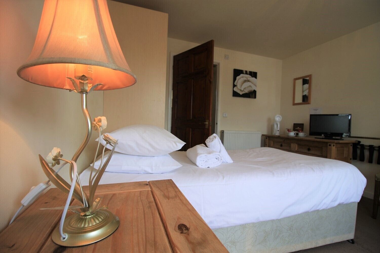 A hotel room with a bed, a wooden nightstand with a lamp, a television, and a radiator.