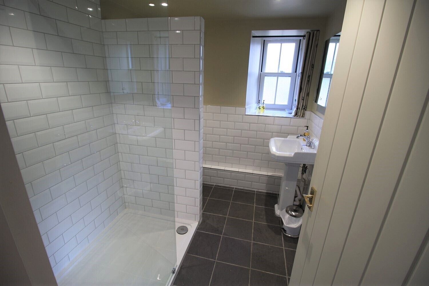 Un baño con ducha de vidrio, un lavamanos blanco y una ventana sobre el lavamanos.