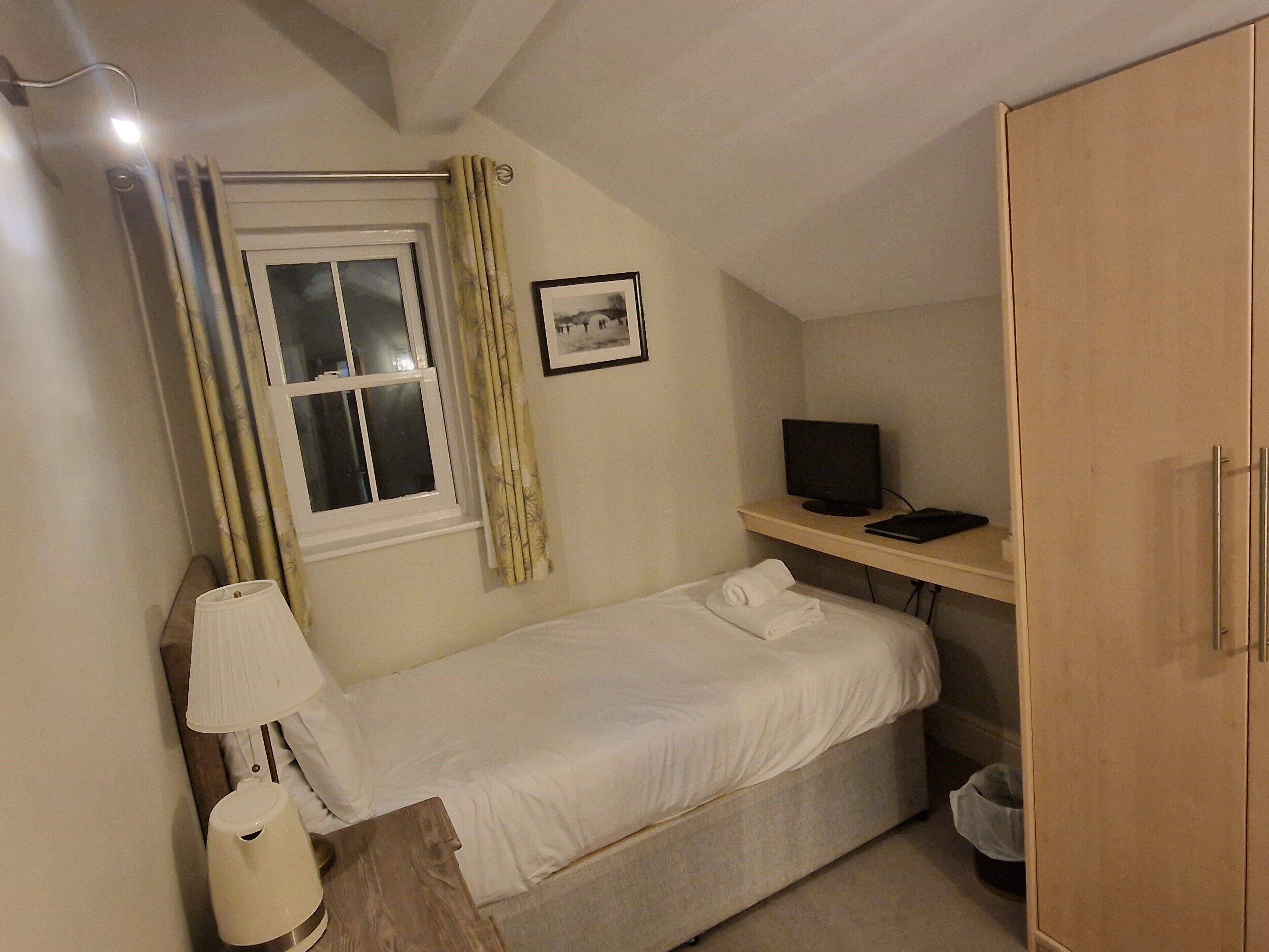A small, single-bed room with a window, a desk with a computer, a lamp, and a wardrobe.