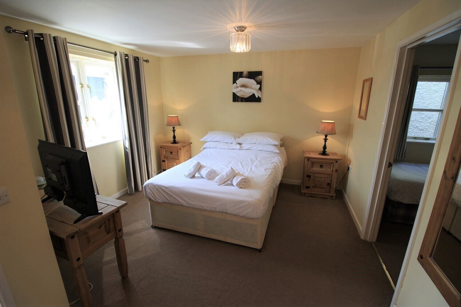 A bedroom with a bed, two bedside tables with lamps, a ceiling light, a wall-mounted picture, and a television on a desk.