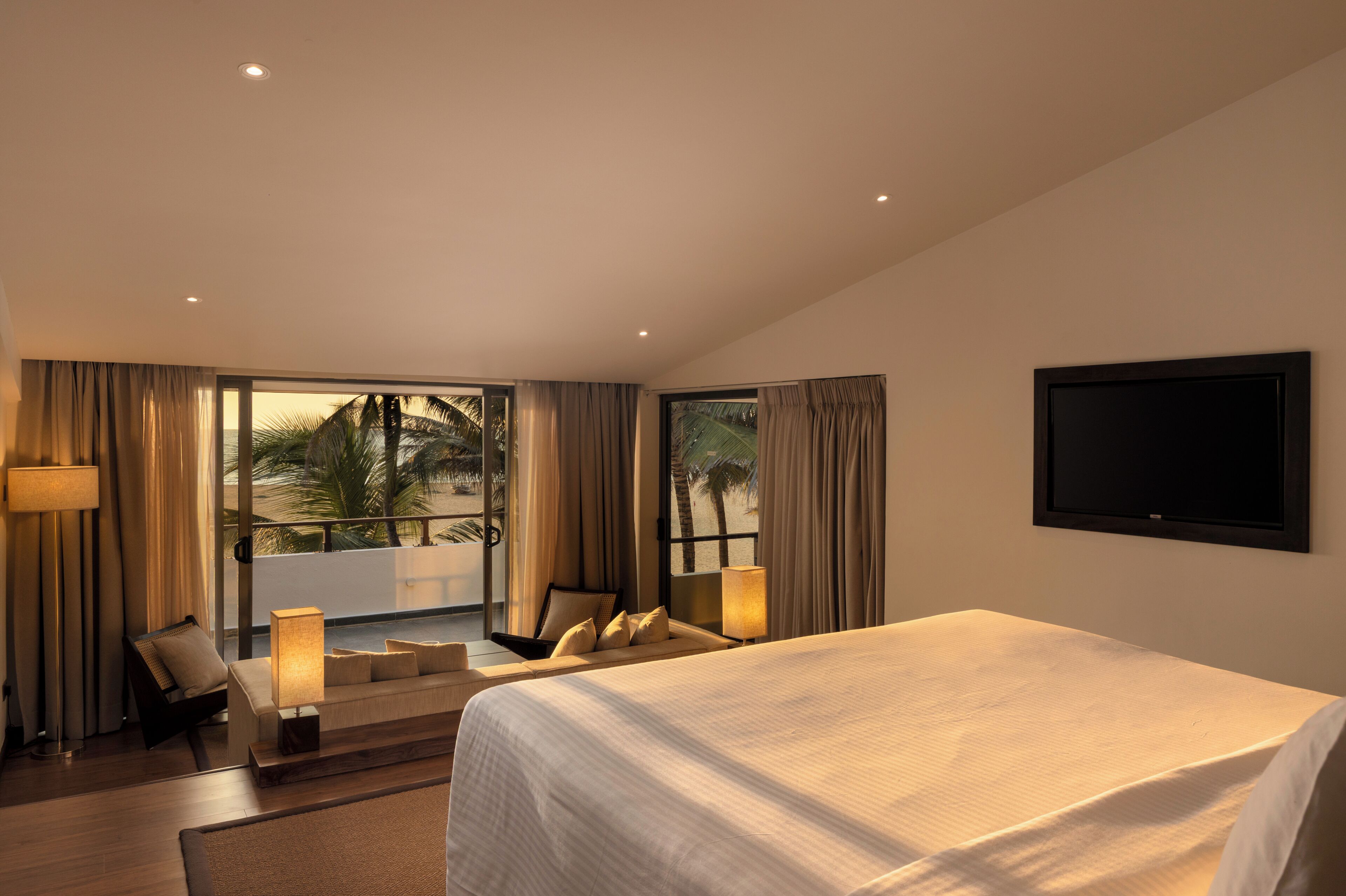 A hotel room with a large bed, a sofa, a chair, a TV, and a balcony with a view of palm trees.