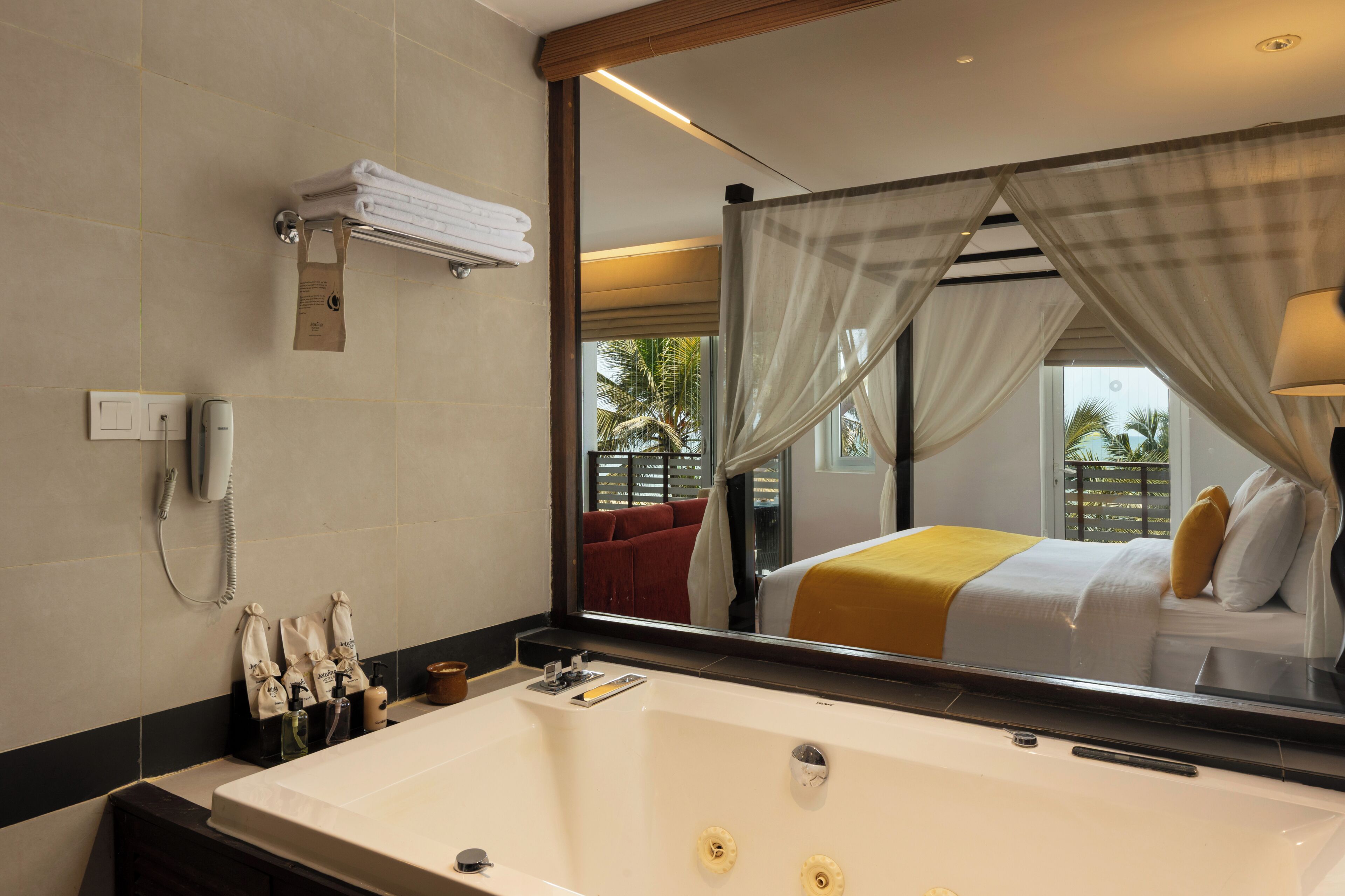 A hotel bathroom with a large bathtub, a telephone, and a mirror reflecting a bed with a yellow blanket.