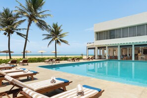 A poolside area with lounge chairs, a pool, palm trees, and a building with large windows.