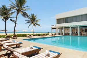 Área à beira da piscina com espreguiçadeiras, piscina, palmeiras e um edifício com grandes janelas.