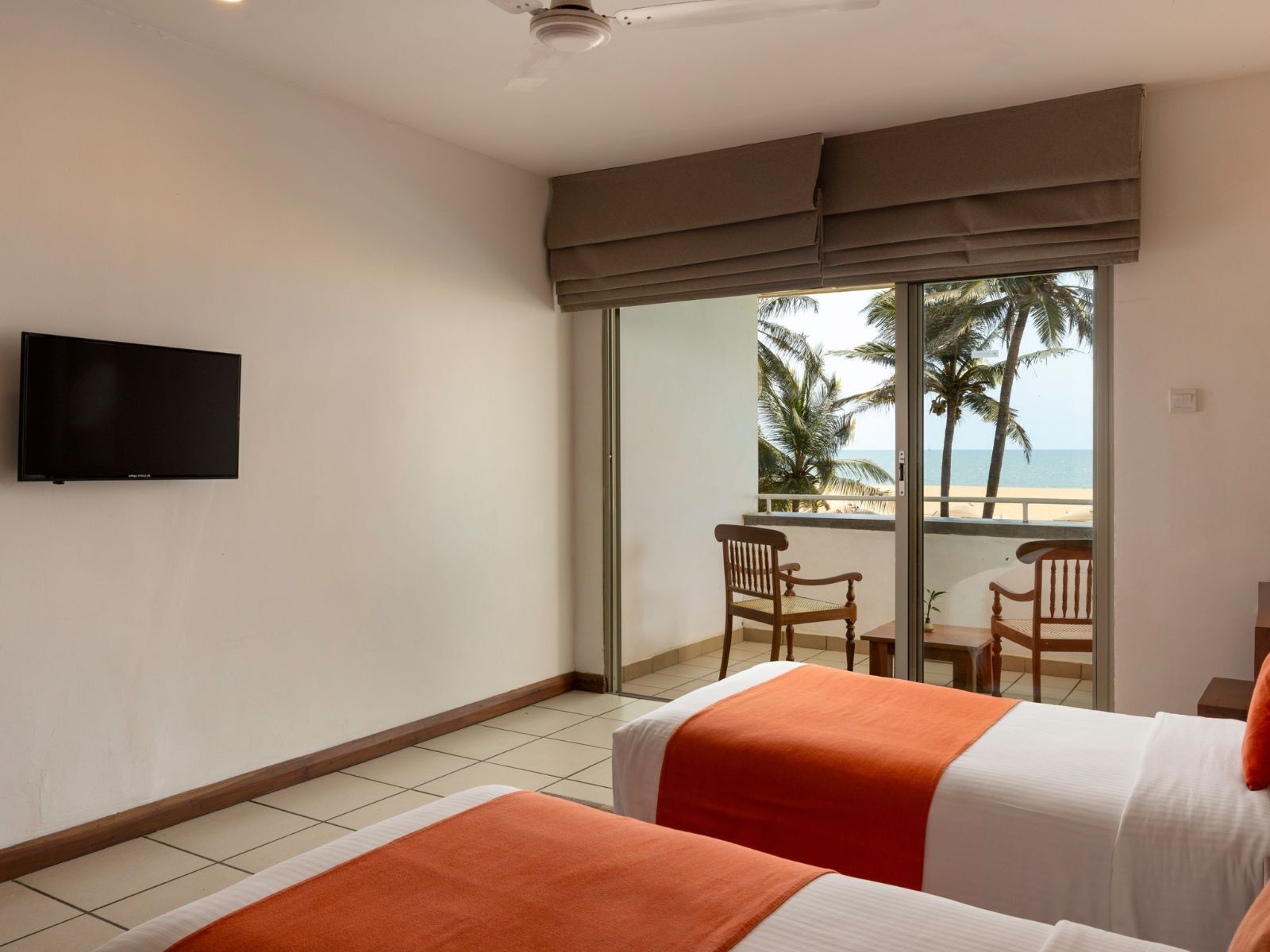 Une chambre d’hôtel avec deux lits, une télévision, un balcon donnant sur la plage et les palmiers, et un ventilateur de plafond.