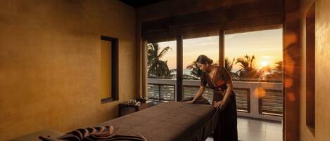 A room with a bed, a window, and a view of palm trees and the sunset.