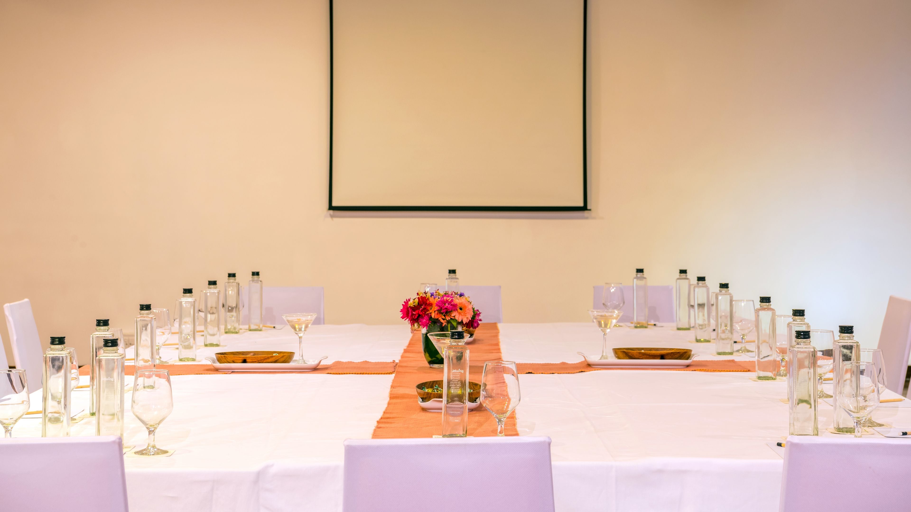 Une salle de conférence avec une longue table préparée pour une réunion, entourée de chaises, et un grand écran fixé au mur.