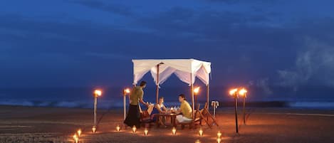 A group of people dining under a canopy on a beach at night, with candles and torches providing light.