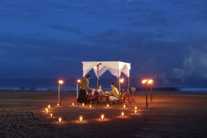 A group of people dining under a canopy on a beach at night, with candles and torches providing light.